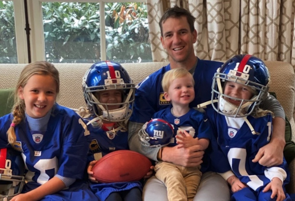Eli Manning on the couch with his four children, all smiling at the camera and wearing his Giants jersey.
