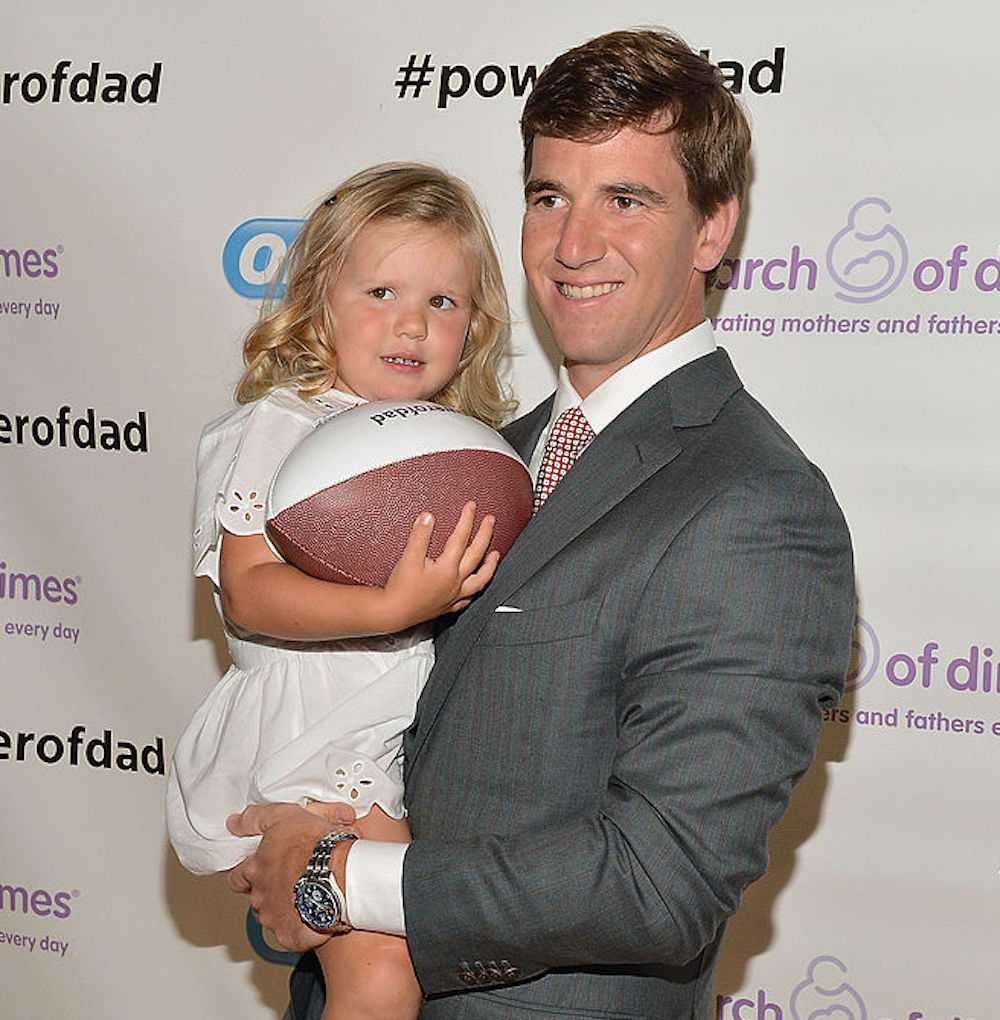 Eli Manning holding daughter Ava Francis while she holds a football and smiles for cameras.