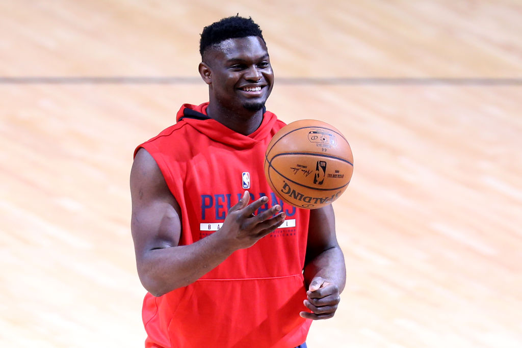 Zion Williamson wearing a red cut off Pelicans NBA team hoodie, tossing a basketball up and smiling, looking fit and not fat.