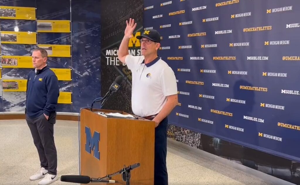 Jim Harbaugh at podium with his hand up