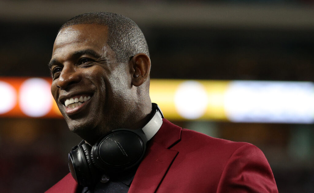 Deion Sanders smiling with jacket and headphones.