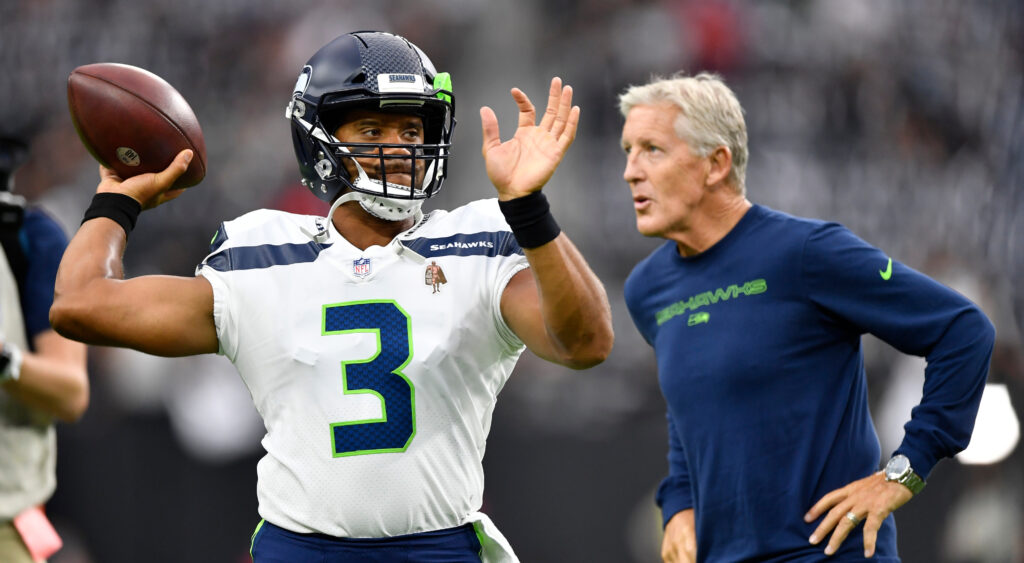 Russell Wilson throwing a football with Pete Carroll in the background