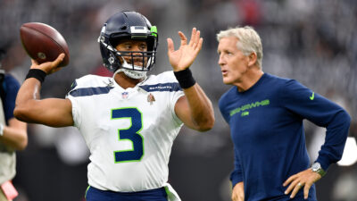 Russell Wilson throwing a football with Pete Carroll in the background