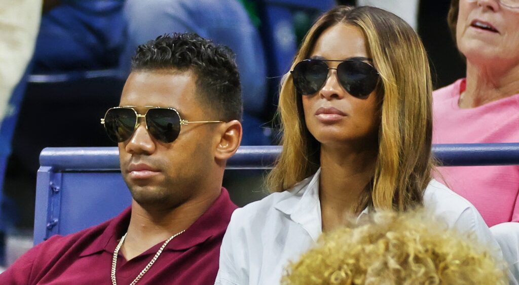 Russell Wilson in sun glasses sitting next to Ciara