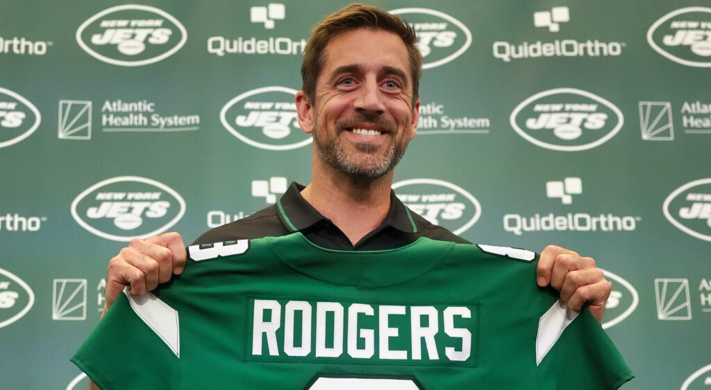 Aaron Rodgers smiling holding Jets jersey.