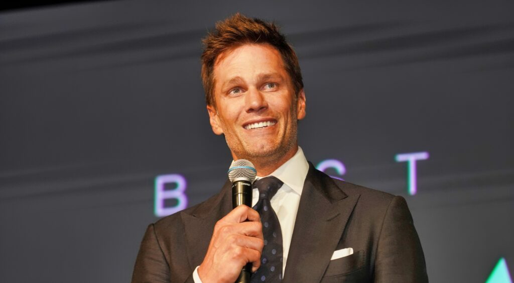 Tom Brady speaking at event with microphone in hand.